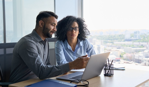 Young man and woman sit at a desk in a high rise building going over something on a laptop