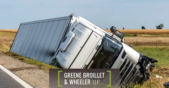 After an accident, a tractor-trailer lays on its side in a field next to a highway on a sunny day