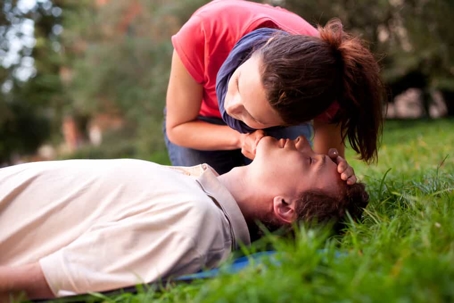 Young woman checks a young man's pulse who is laying face up on the grass in a park during the day
