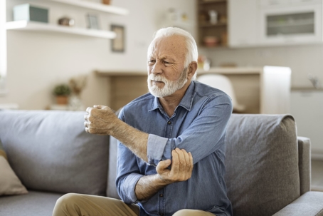 Senior man sits on couch and clutches his elbow in pain