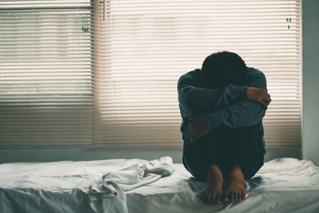 Person sits on a bed holding their legs in sadness